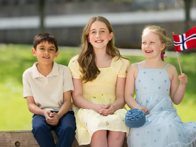 Gutt i hvit piquetskjorte, blå bukse, jente i gul kjole, jente i bå kjole sitter ved siden av hverandre ute.