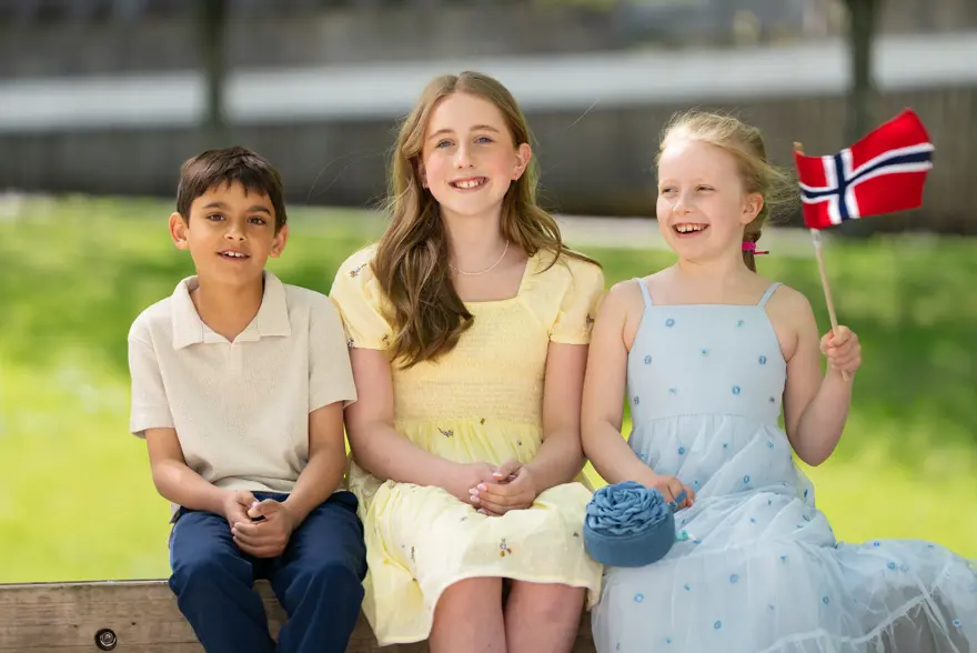 Gutt i hvit piquetskjorte, blå bukse, jente i gul kjole, jente i bå kjole sitter ved siden av hverandre ute.