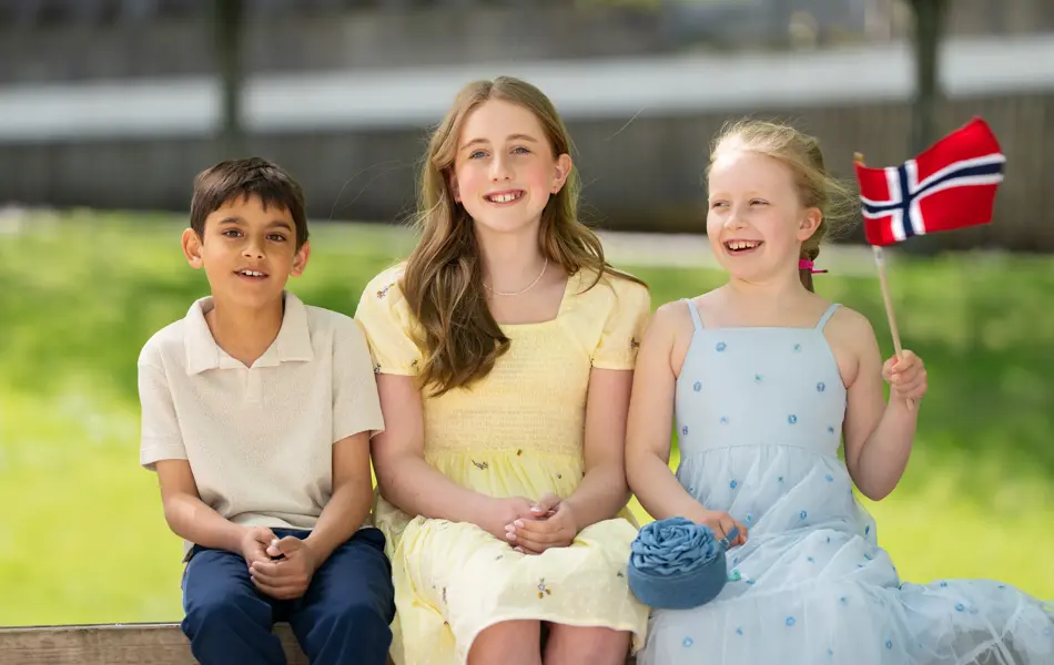 Gutt i hvit piquetskjorte, blå bukse, jente i gul kjole, jente i bå kjole sitter ved siden av hverandre ute.