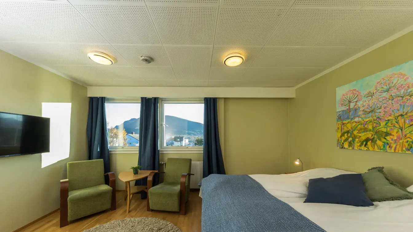 Dobbeltseng med pynteputer, vegghengt TV og to stoler ved vinduet