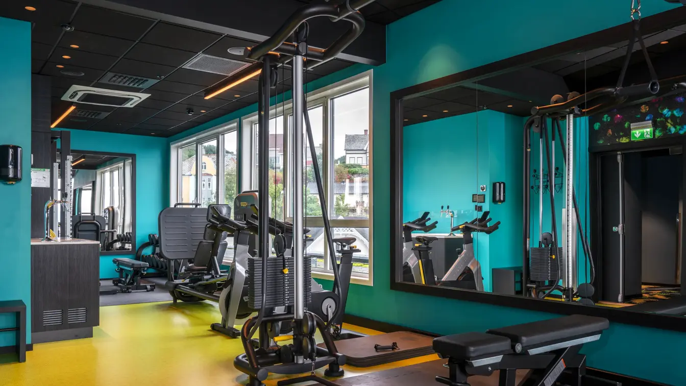 Turkis vegg, stort veggspeil, svart og gult gulv, vinduer fra gulv til tak, diverse treningsapparater. 