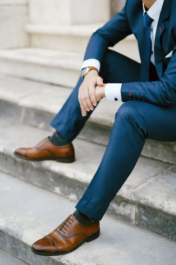 Nærbilde av gutt i blå dress med brune sko, klokke og mansjettknapper, som sitter i trapp. Foto til artikkel om kjøp av konfirmasjonsdress. 