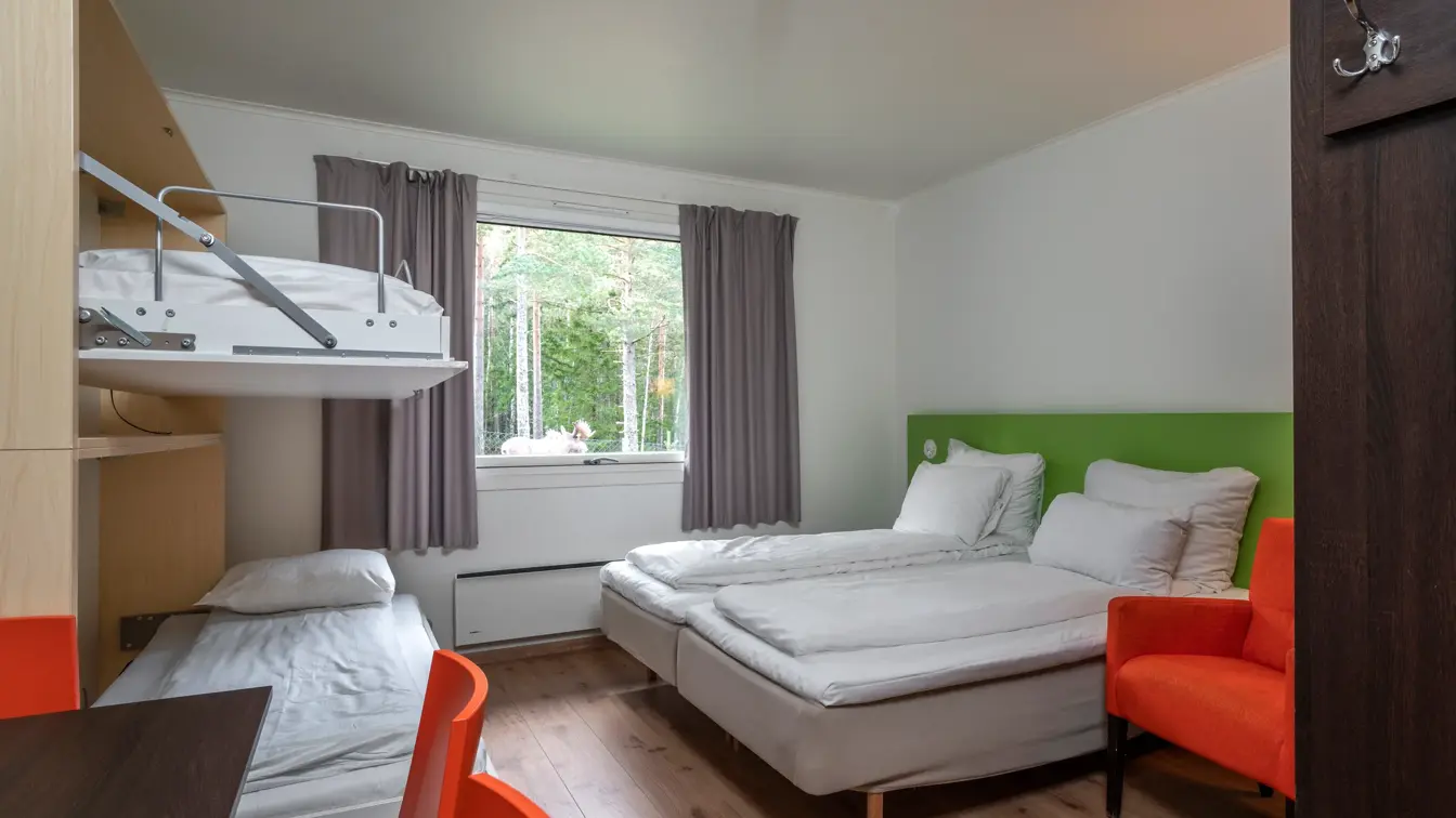 Studioleilighet med dobbeltseng og køyesenger, vindu med grå gardiner, grønn sengegavl, oransje lenestol