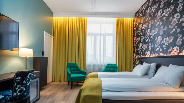 Stort vindu med gule gardiner fra tak til gulv. To grønne lenestoler. Mørk tapet med store lyserosa roser. To enkeltsenger med gule sengetepper. Mørkt skrivebord med veggehengt TV og mønstrete stol. I standard twin rom på Thon Hotel Høyers i Skien.