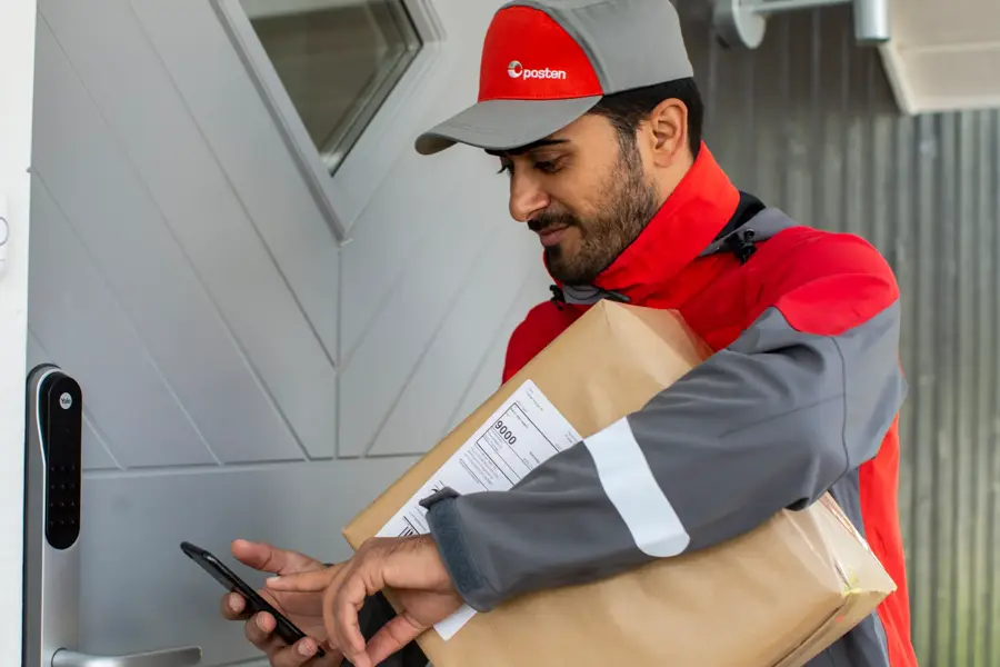 Mann iført arbeidsklær fra Posten står utenfor en dør med en pakke under armen som skal leveres