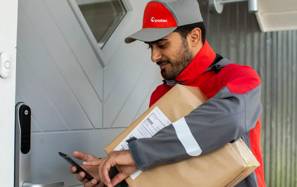 Mann iført arbeidsklær fra Posten står utenfor en dør med en pakke under armen som skal leveres