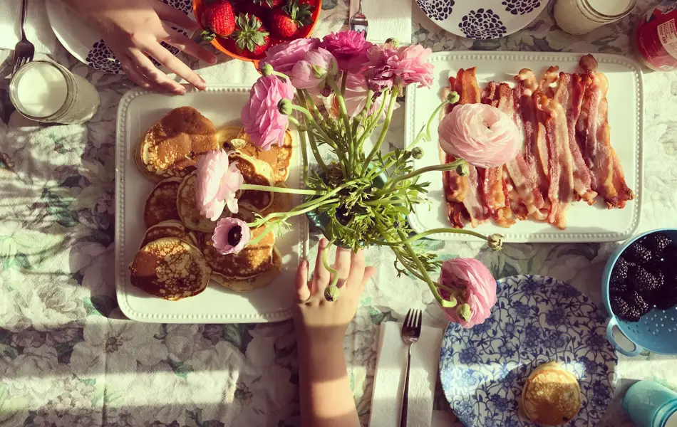 Bird's eye view of a breakfast table set for three with colorful plates, tablecloth, pancakes, fruit and bacon. A glimpse of three kids hands resting on the table. Childhood. Friendship.
