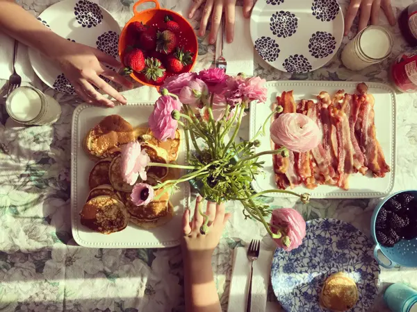 Bird's eye view of a breakfast table set for three with colorful plates, tablecloth, pancakes, fruit and bacon. A glimpse of three kids hands resting on the table. Childhood. Friendship.