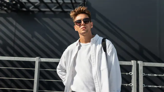 Urban young man hipster in trendy white shirt in fashionable sunglasses with a cloth black bag posing on a sunny day near a gray building outdoors in the city. Trendy guy. Youth summer style.