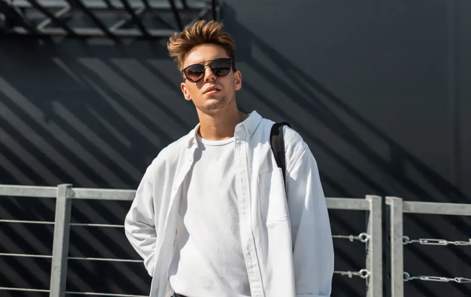 Urban young man hipster in trendy white shirt in fashionable sunglasses with a cloth black bag posing on a sunny day near a gray building outdoors in the city. Trendy guy. Youth summer style.