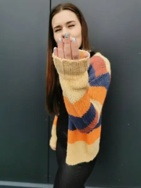 Jente i skinntights og fargerik stripete cardigan holder opp den ene hånda foran seg for å vise frem neglelakken. Neglelakken er sølvfarget, og det kan se ut som den glitrer. 