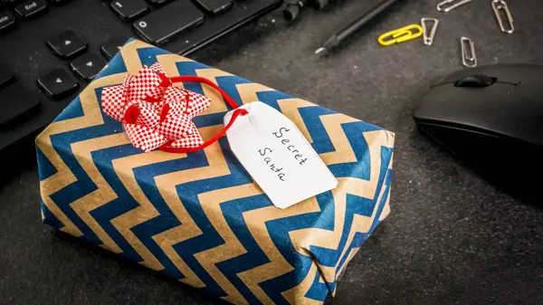 Office Christmas celebration concept, the idea of sharing gifts secret Santa. Keyboard, mouse, notebook, pens, pencils, Christmas gift. Black office table, copy space