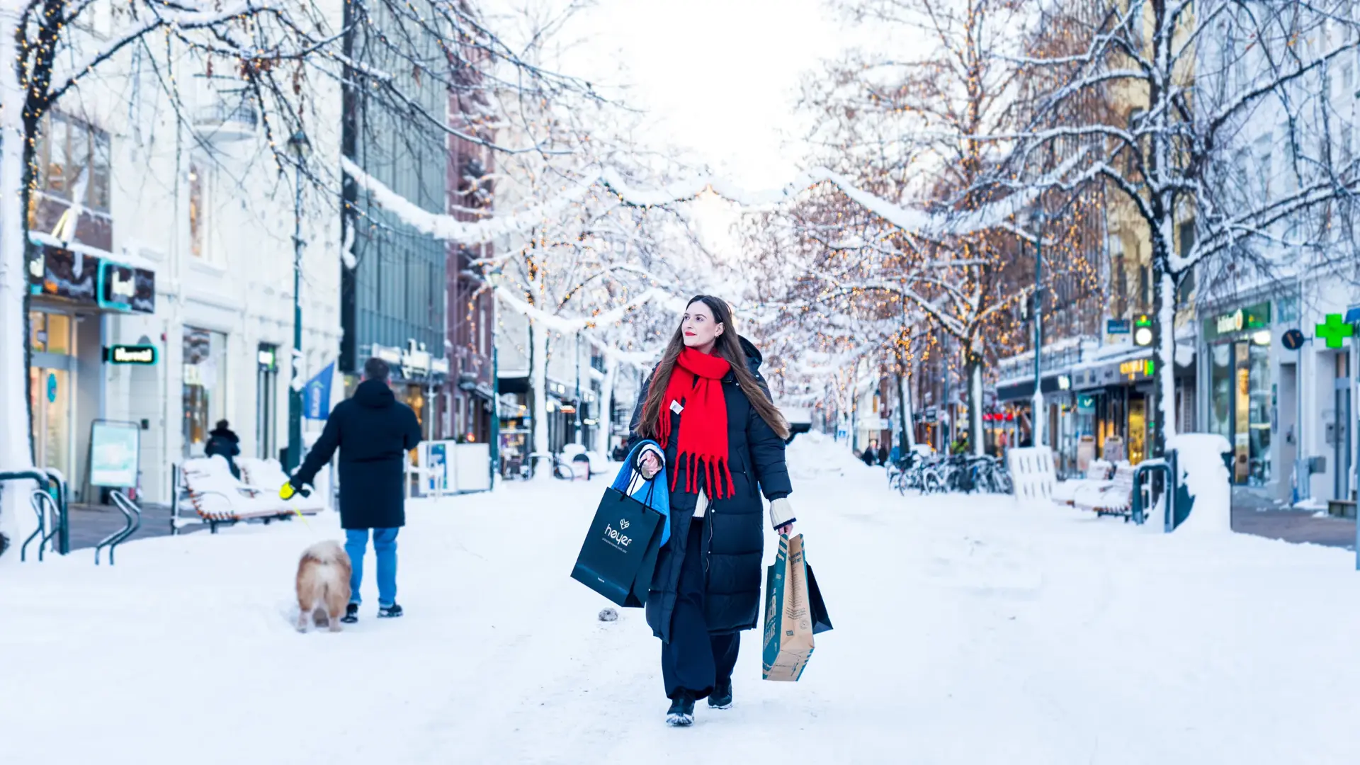 Kvinne med handleposer går i en snødekt Nordre gate i Trondheim. Det er vinter, med julelys og butikker på begge sider.