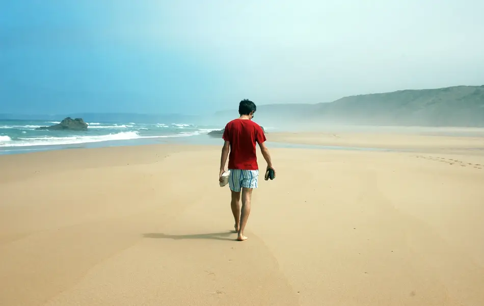 Mann på øde strand med rød t-skjorte og stripete shorts. Bølgeskvulp og sand.
