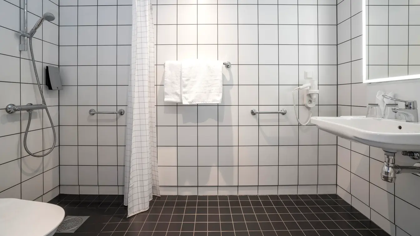 Dusj, servant, speil med led lys rundt, hvite fliser på vegger og mørke på gulv i romslig bad i handicap rom på Thon Hotel Kirkenes