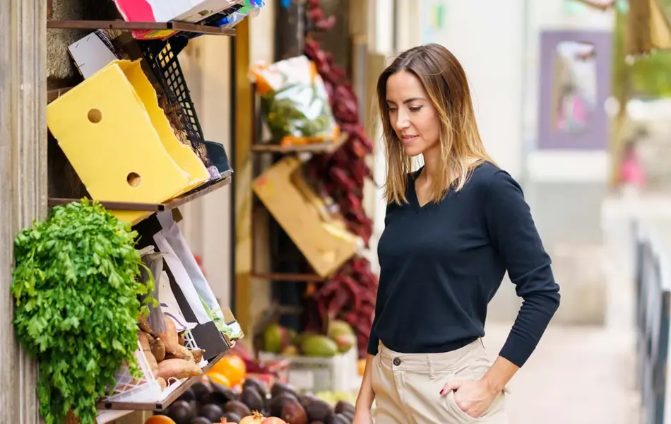 Dame iført blå, tettsittende genser og lys cargobukse ser på grønnsaker utenfor en butikk.