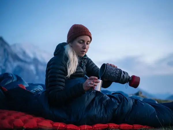 Kvinne som sitter på liggeunderlag inni sovepose ute i snøen og heller varm drikke fra termos