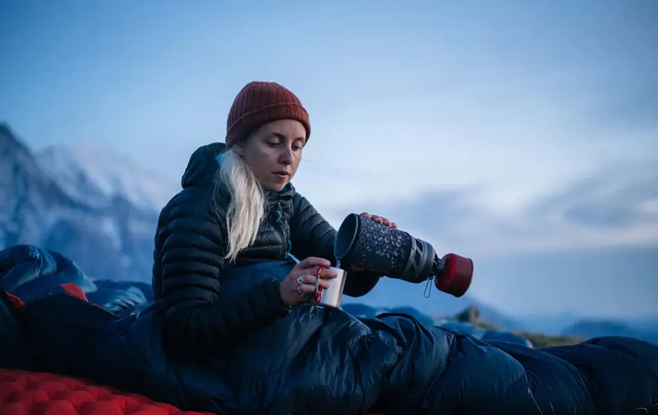 Kvinne som sitter på liggeunderlag inni sovepose ute i snøen og heller varm drikke fra termos