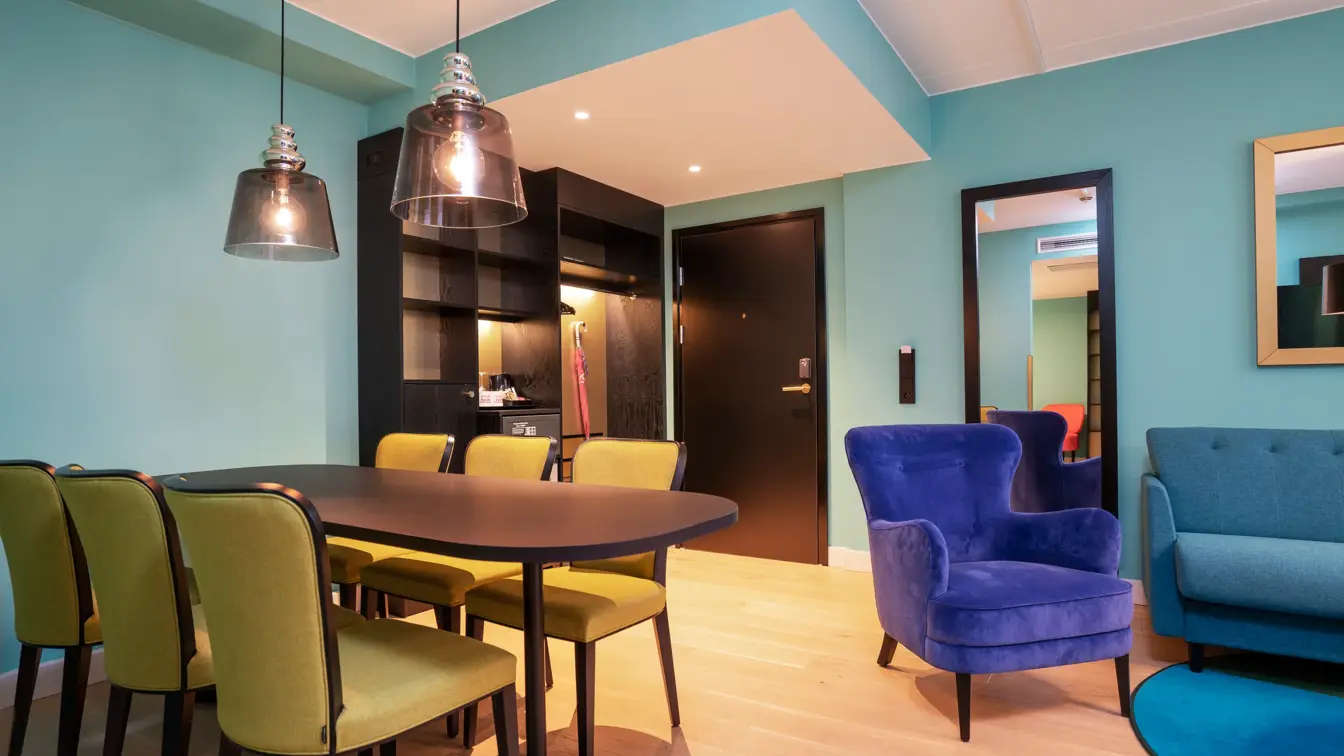 Turkis tapet, spisebord med gule stoler og mørkt bord, stort speil, sittegruppe med turkis siofa og blå stol, i junior suite på Thon Hotel Storo i Oslo