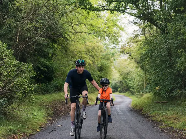Mann og gutt sykler mot kamera på landevei med frodig skog rundt seg. Foto til artikkel om gaver til pappatyper på farsdag. 