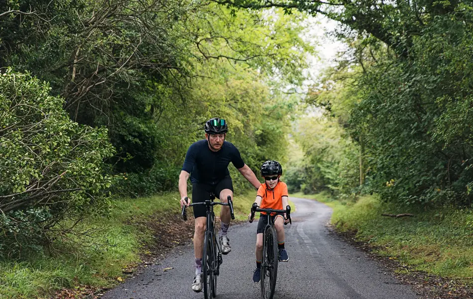 Mann og gutt sykler mot kamera på landevei med frodig skog rundt seg. Foto til artikkel om gaver til pappatyper på farsdag. 