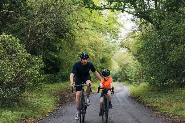Mann og gutt sykler mot kamera på landevei med frodig skog rundt seg. Foto til artikkel om gaver til pappatyper på farsdag. 