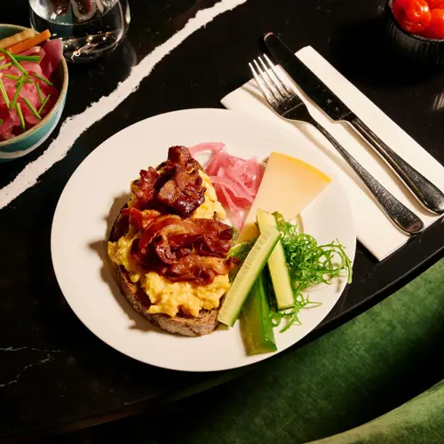 En frokosttallerken med en skive grovbrød toppet med eggerøre og sprøstekt bacon. Ved siden av ligger syltet rødløk, ost, agurk og salat. På bordet står også en skål med grønnsaker og en med små tomater.