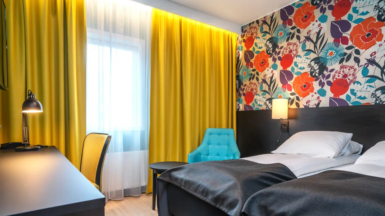 Blomstrete tapet, et stort vindu fra gulv til tak, gule gardiner, parkettgulv, to enkle senger, svart skrivebord, en gul stol og en blå stol. 