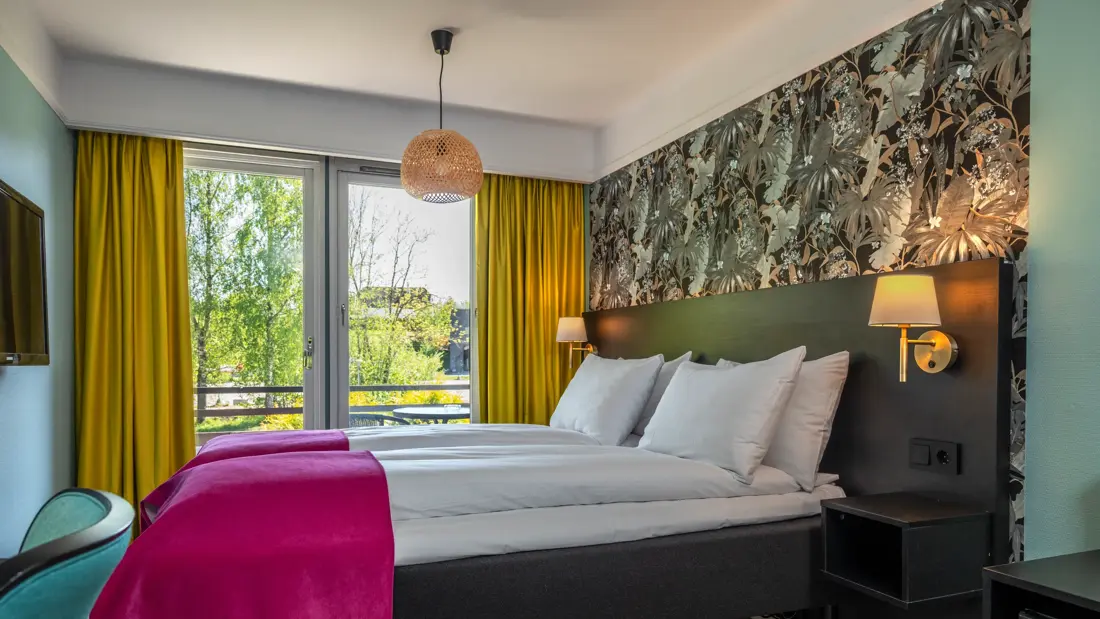 Standardrom med twin senger, mønstrete tapet, store vinduer med gule gardiner, rosa sengeteppe, parkett og vegghengt TV