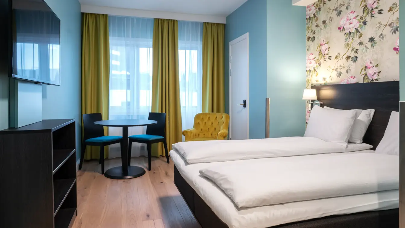 Blomstrete tapetvegg, blå vegg, dobbeltseng, vindu fra gulv til tak, TV, gul stol, salongbord med to stoler, 