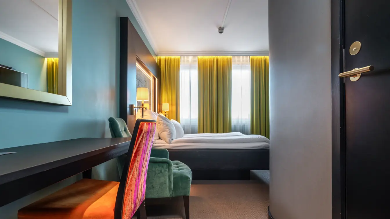 Dobbeltrom med dobbeltseng, gule gardiner, turkis vegg, skrivebord med oransje stol.