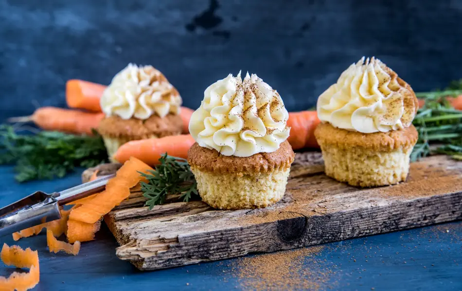 Tre gulerotmuffins med topping på trefjøl omringet av gulerøtter og grønt