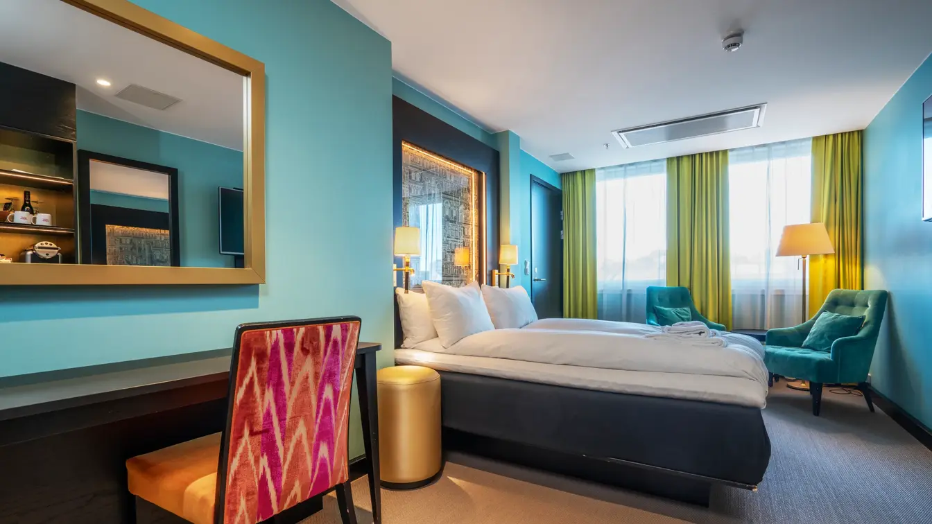 Dobbeltrom med dobbeltseng, turkise vegger, skrivebord med oransje stol, grønne lenestoler og gule gardiner.