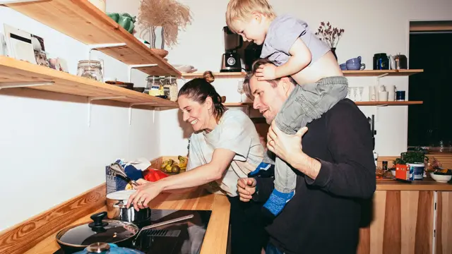 Familie med mor, far og sønn holder på på kjøkken med trebenk og fylte trehyller 