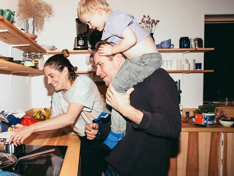 Familie med mor, far og sønn holder på på kjøkken med trebenk og fylte trehyller 