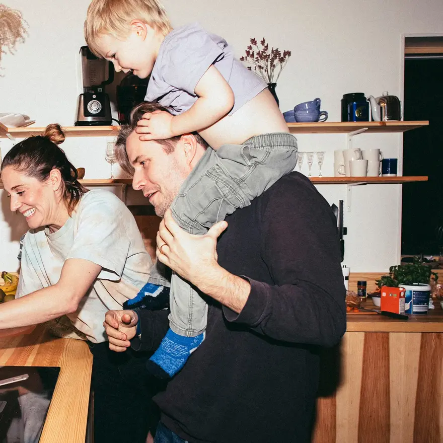 Familie med mor, far og sønn holder på på kjøkken med trebenk og fylte trehyller 