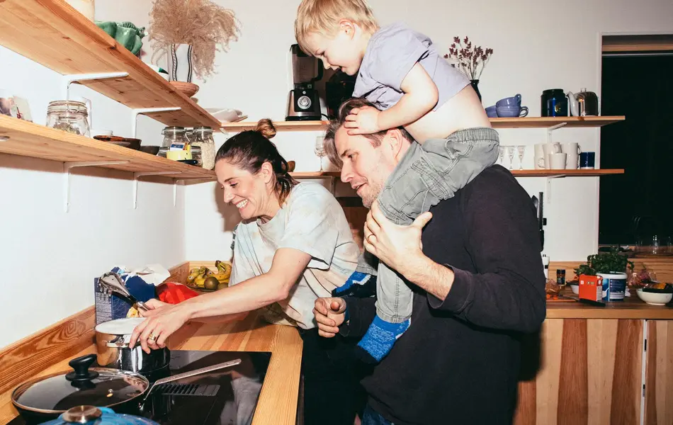 Familie med mor, far og sønn holder på på kjøkken med trebenk og fylte trehyller 