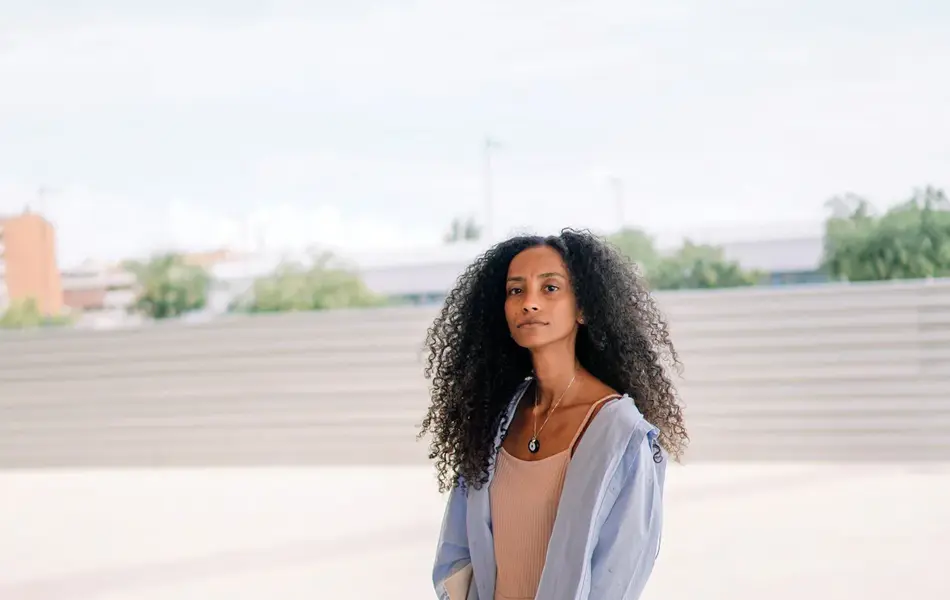 Kvinne i helfigur, med rosa kjole og lyseblå overdel, står på åpen plass i byrom og ser i retning kamera.