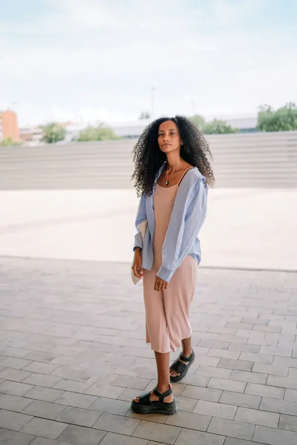 Kvinne i helfigur, med rosa kjole og lyseblå overdel, står på åpen plass i byrom og ser i retning kamera.