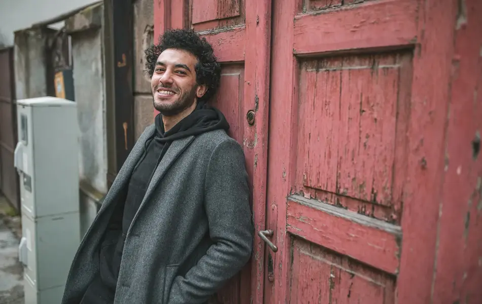 Mann med grå frakk og sort hettegenser lener seg inntil en dør utendørs 