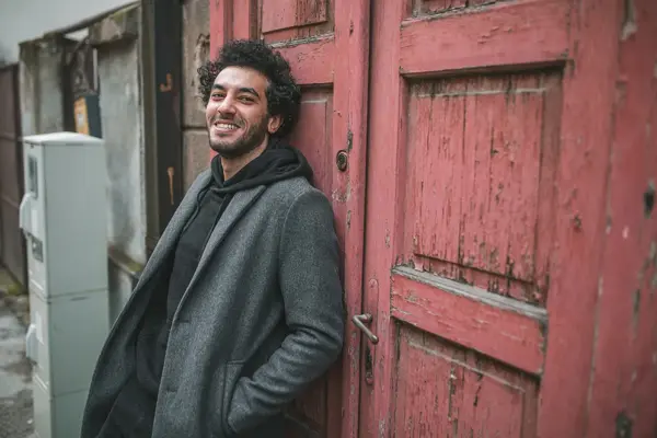 Mann med grå frakk og sort hettegenser lener seg inntil en dør utendørs 