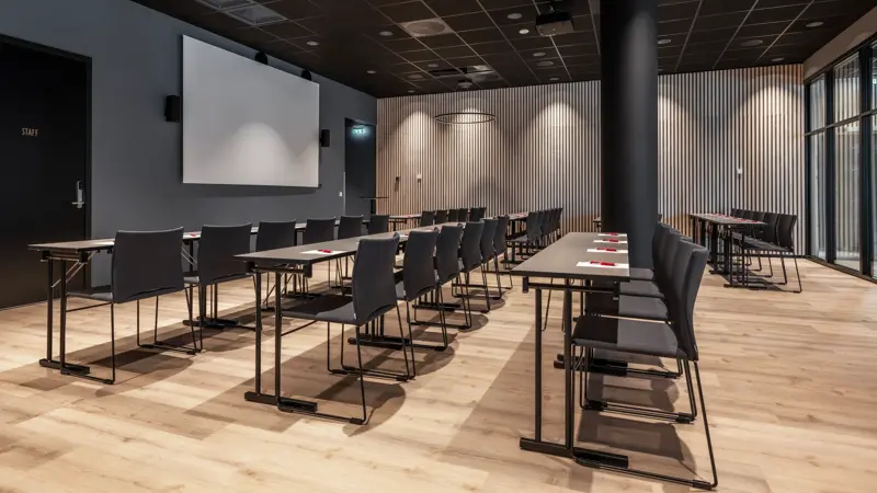 Konferanserom med klasserom oppsett, stort lerret, svart tak, og parkett.