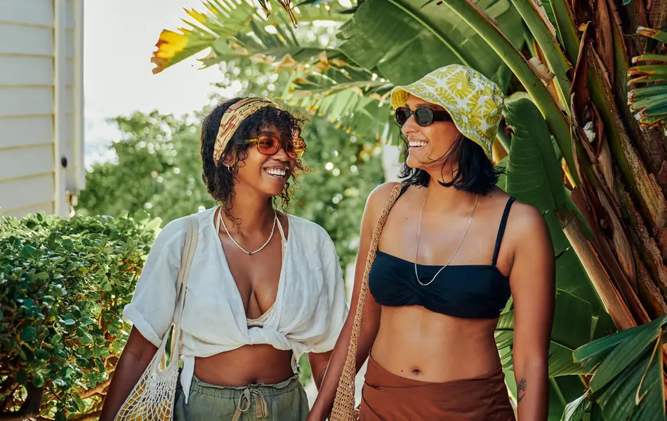 To damer med solbriller, bøttehatt, hodetørke og fiskenettsvesker smiler mens de går foran palmetrær 