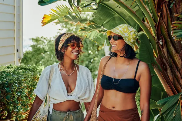 To damer med solbriller, bøttehatt, hodetørke og fiskenettsvesker smiler mens de går foran palmetrær 