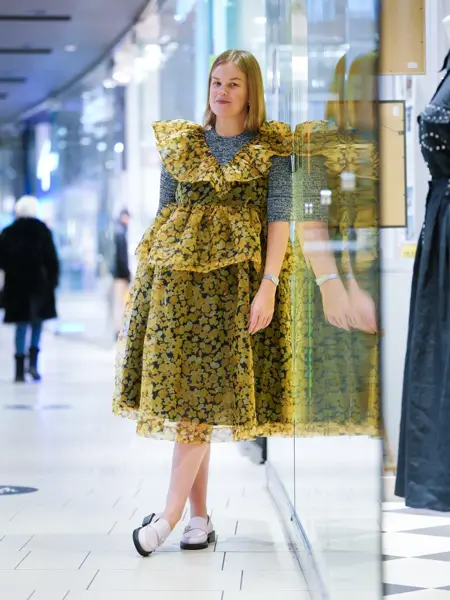 Jente som står og lener seg til en vegg med gul kjole og grå genser under