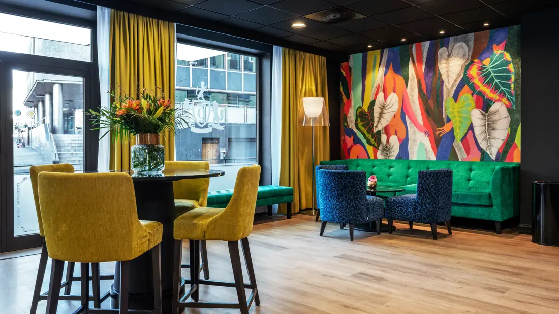 Lobbyområde med fargerike møbler, både sofa, stoler og sittegruppe. Fargerik tapet, gule gardiner og store vinduer ut mot gaten.