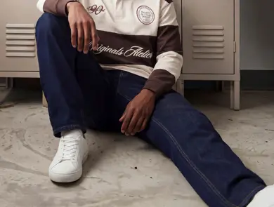 En mann i hvit og brun genser og blå jeans sitter på bakken og lener seg mot noen skap.