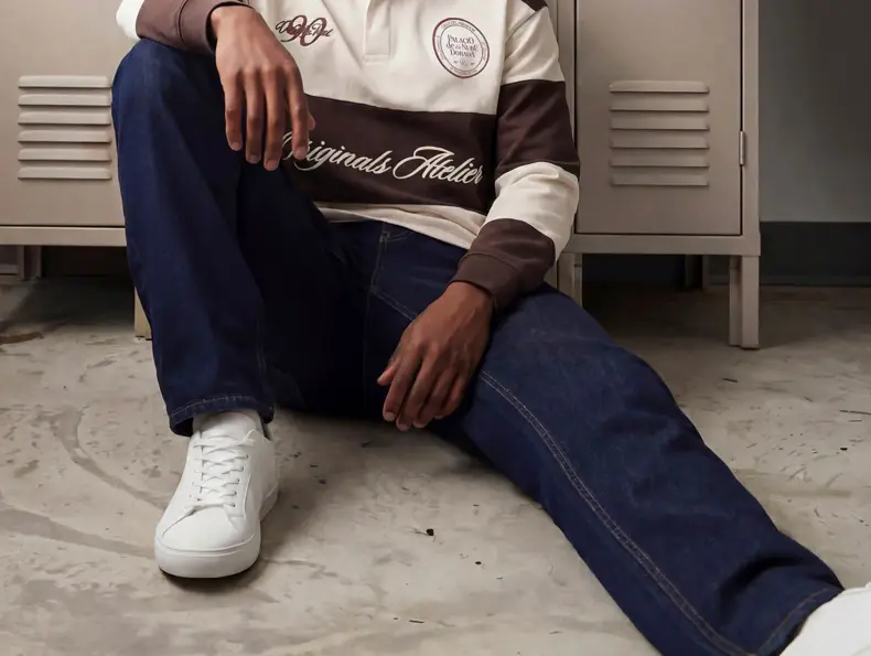 En mann i hvit og brun genser og blå jeans sitter på bakken og lener seg mot noen skap.