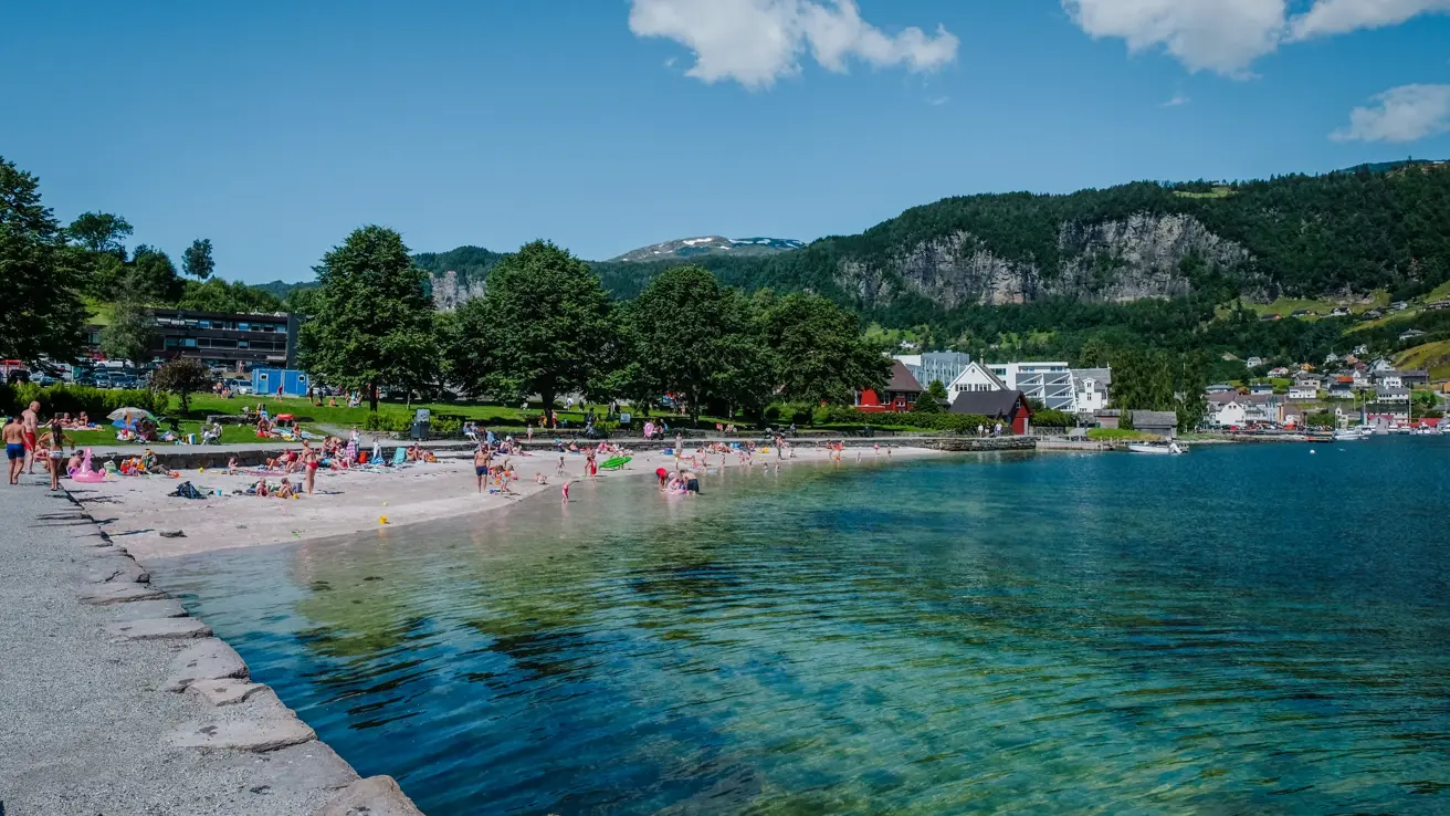 Bystranda i Kristiansand en sommerdag. Turkis vann og mange badegjester på stranda og i vannet.