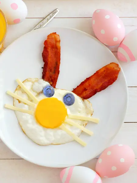 Easter breakfast with cute bunny face made of egg and bacon. Table scene, above view over a white wood background.